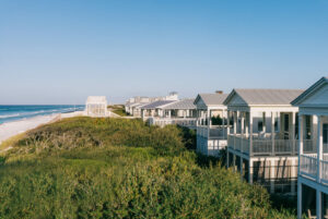 Coastal beach houses overlooking grassy dunes with the ocean and shoreline visible under a clear blue sky.